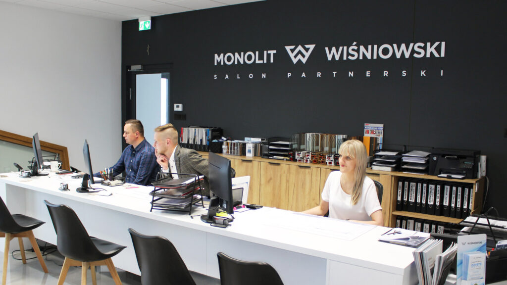 Jesteśmy salonem partnerskim Wiśniowski. Zdjęcie na którym są widoczni pracownicy salonu firmowego Monolit, Aneta, Maciej i Krzysztof, siedzący przy biurku.